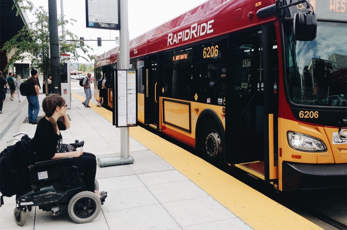 A lady in a wheelchair is waiting to board a bus at a stop with a raised kerb