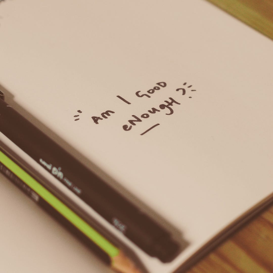 “Am I Good Enough?” is written in a notebook on a desk. 