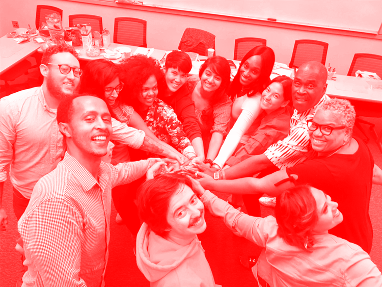 A photo with a monochrome red filter, portraying ten people in a huddle, with their hands together, smiling and looking up at the camera.