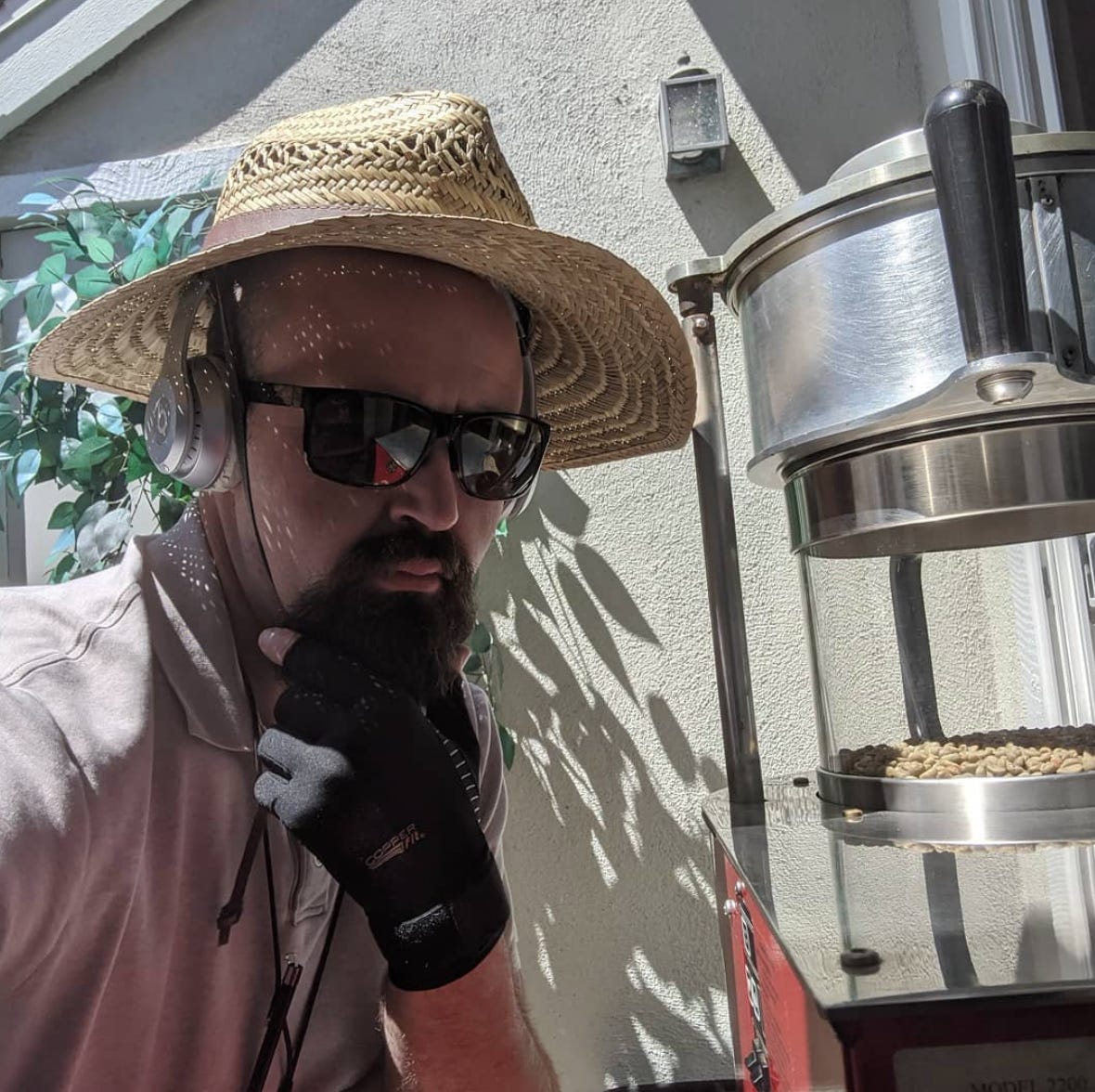 Stephen Crocker caressing his goatee and posing next to his sonos fresco coffee roaster. He's wearing a floppy sun hat, headphones and black sunglasses.