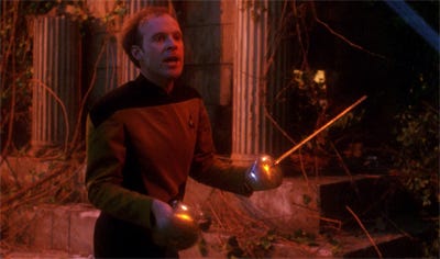 This image shows Lieutenant Reginald Barclay (played by Dwight Schultz) holding an epee (or fencing sword) in each hand while standing in a holographic reproduction of a Grecian temple in Star Trek: The Next Generation's Season 3 episode "Hollow Pursuits."