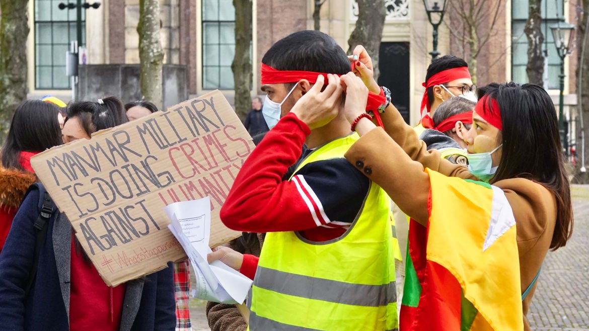 Cover Photo: A photograph of protestors. A woman is tying a red bandanna around a man's head, while he holds a sign that says in all capitals: " Myanmar military is doing crimes against humanity."