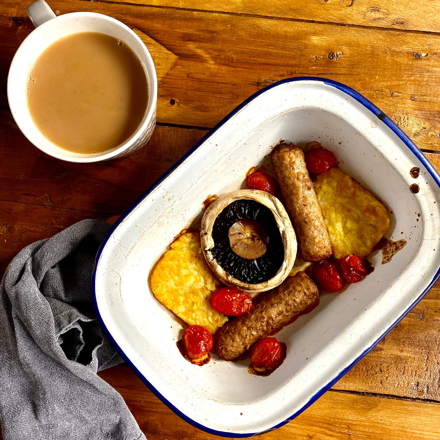 Oven dish with hash browns, mushroom, tomatoes and veggie sausages, a mug of tea to the side