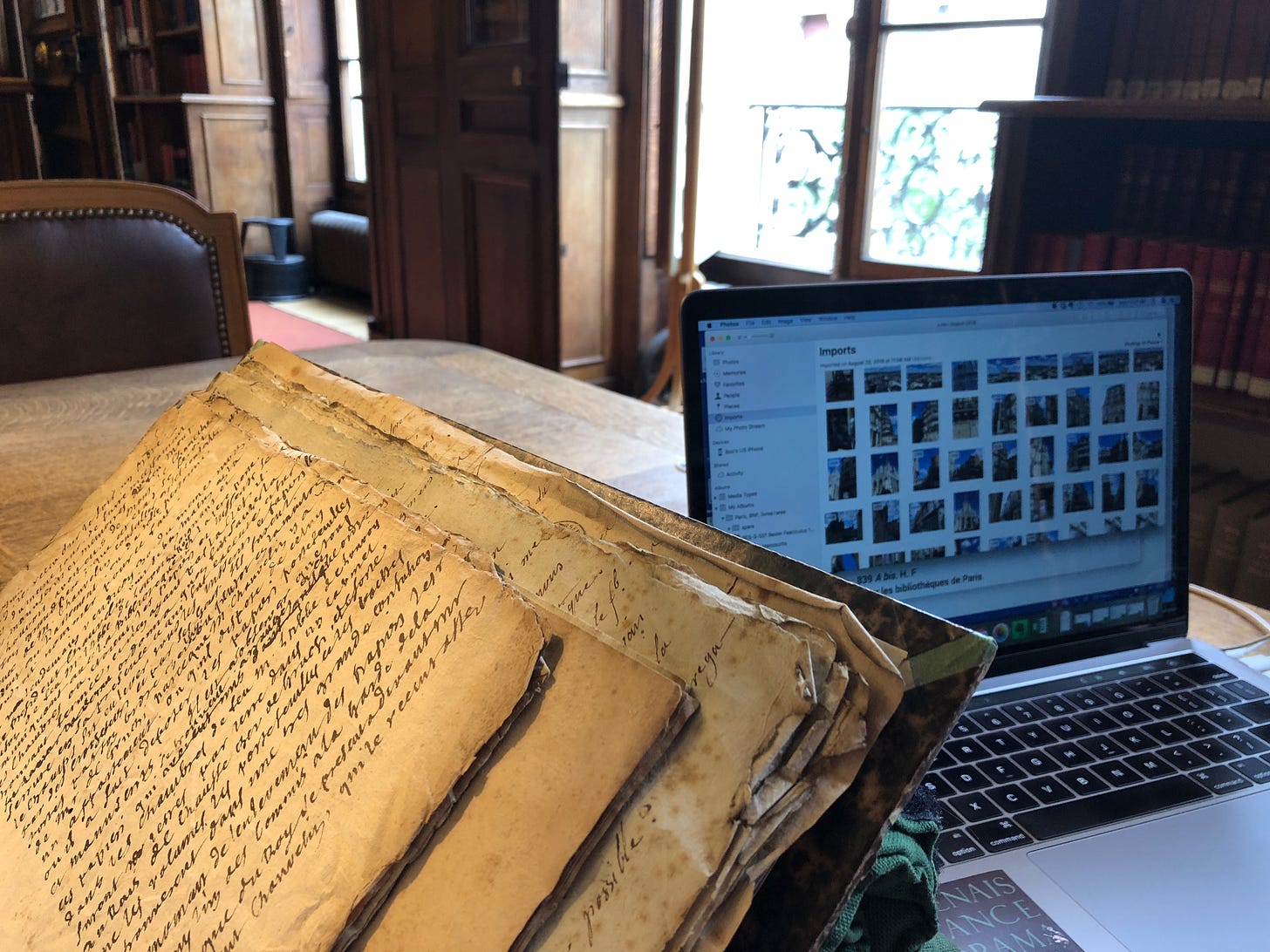 A Macbook showing the Photo program sits on a wooden desk with a manuscript volume in wood-panelled room with large windows.