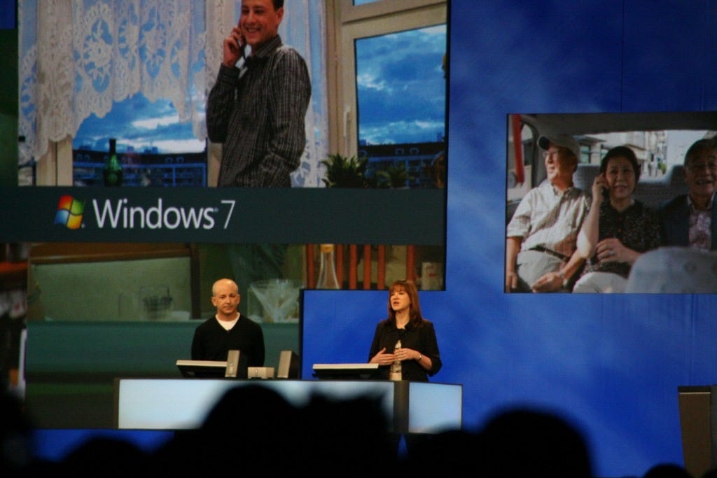 Stage backdrop showing large photos of people using Windows 7. On stage are Steven Sinofsky and Julie Larson Green while Julie does a demo.