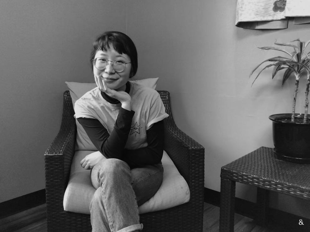Black and white photo of Tiffany sitting on a chair posing with her hand under her chin, smiling.