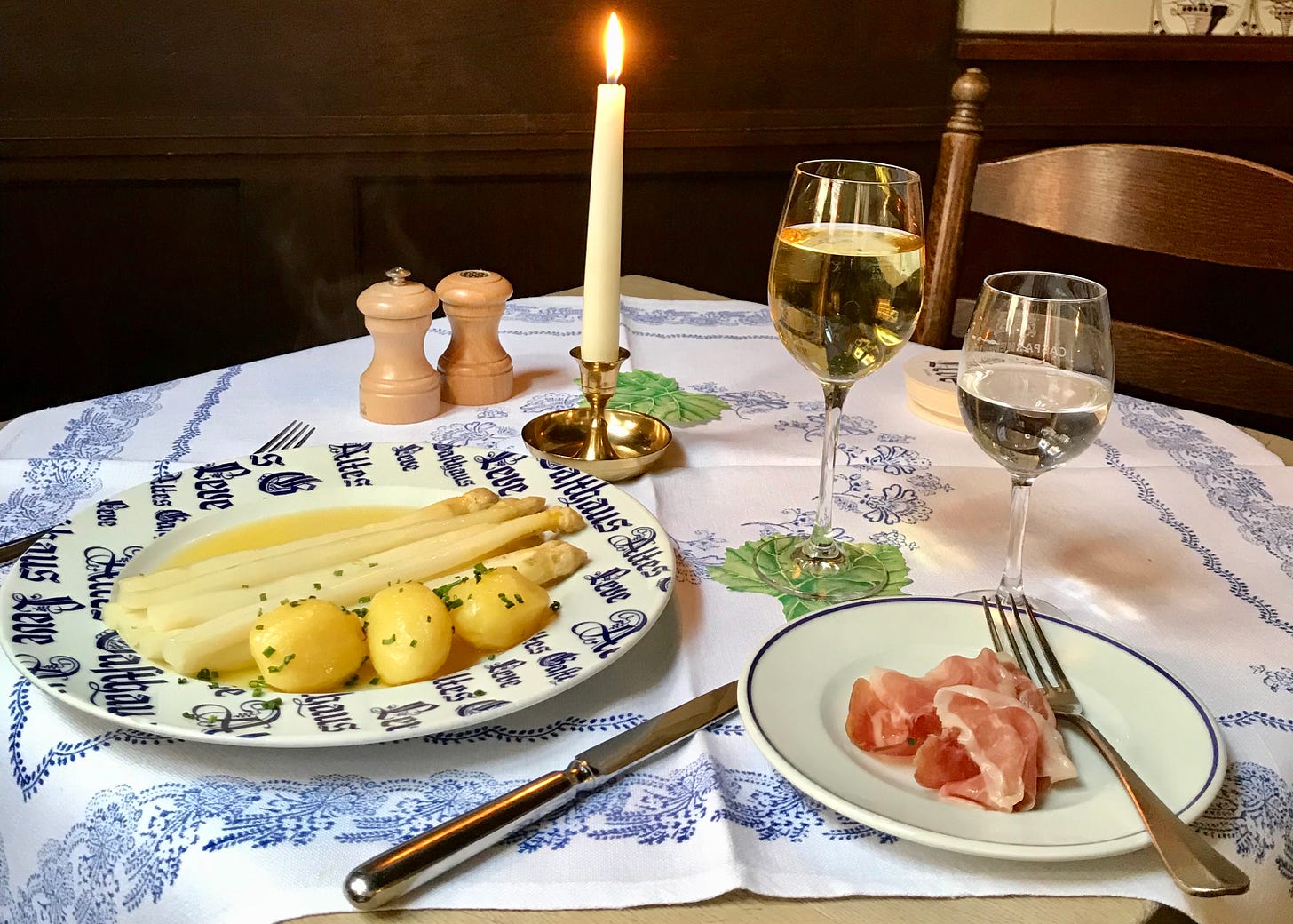 Plate of white asparagus with butter and ham, and a side plate of ham, plus wine glass and water glass, candle and salt and pepper on a blue and white tablecloth