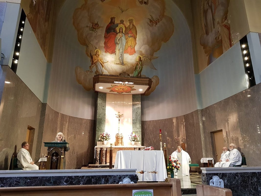 En kyrkinteriör med en målning av helgon i himlen på väggen ovanför altaret och fyra vitklädda präster sittande vid väggarna. En kvinna står vid läspulpeten och läser.
