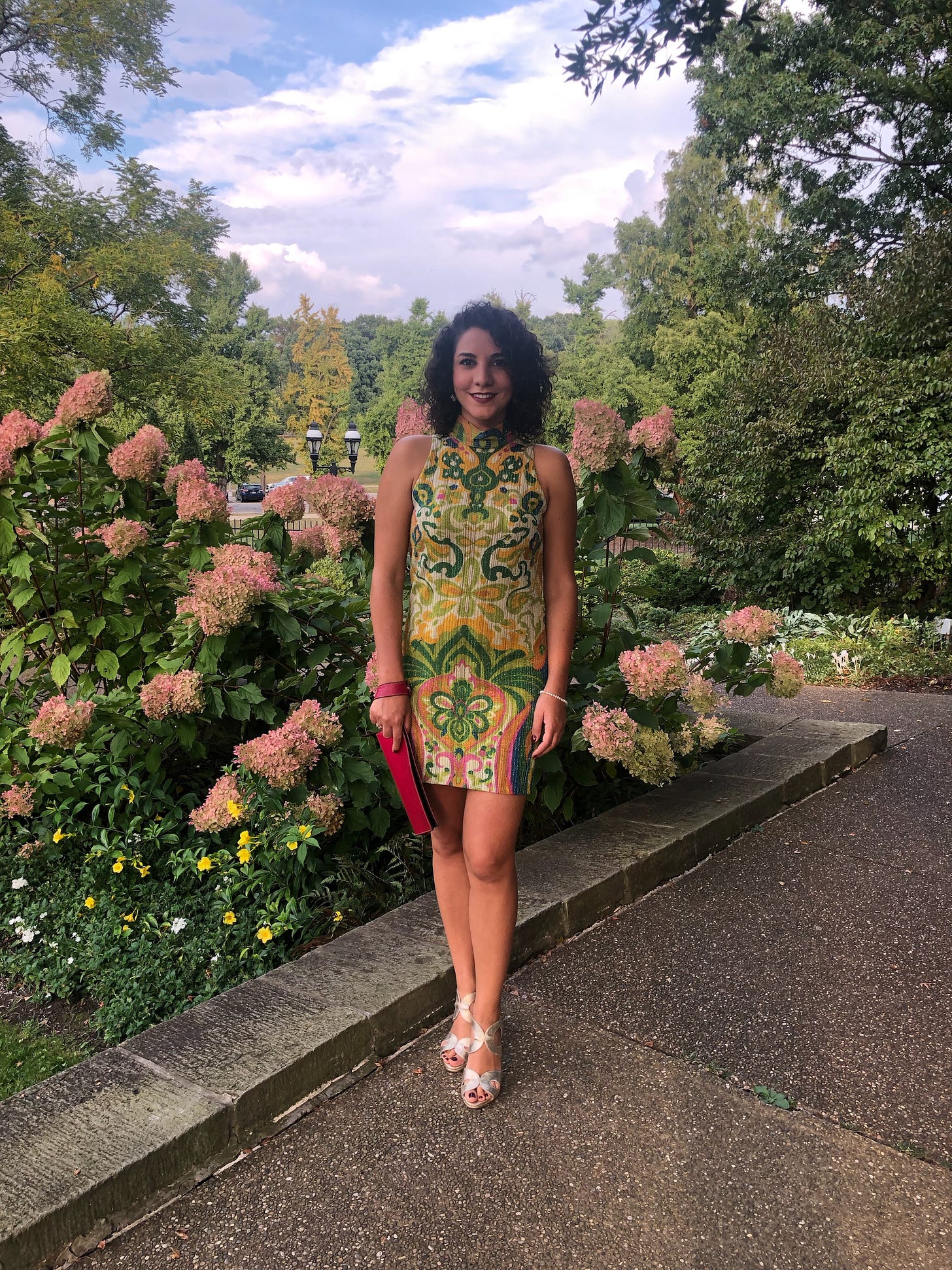 Elda stands in a park on a clear day in front of a pink hydrangea bush. Elda is smiling, her hair black and curly, and she wears a 60s pink and green mini dress and high tan heels