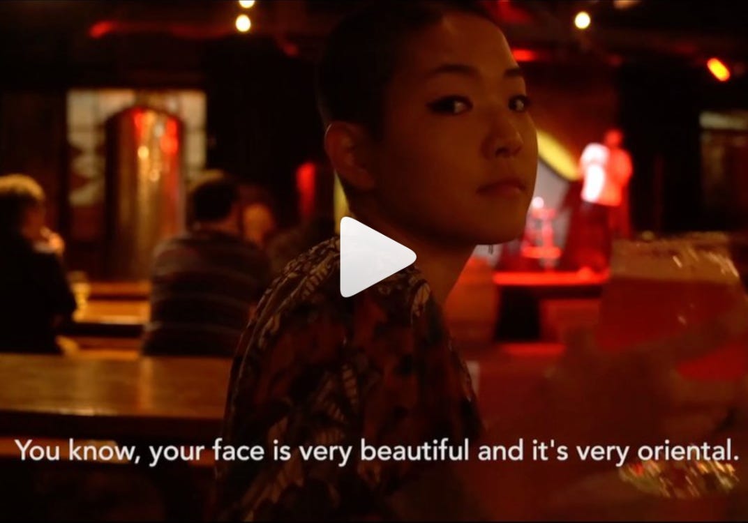 Kyoko Takenaka is pictured, staring at the camera in a bar, with subtitles on the screen that read "you know, your face is very beautiful and it's very oriental." A play button is overlaid on top of the image.