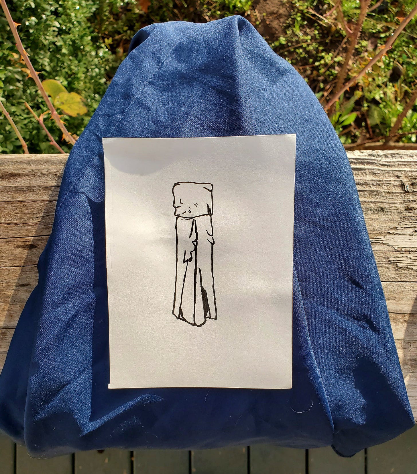 A black and white ink sketch of a strapless, flowy dress on a piece of navy blue fabric, taken outside on a white porch against a green bush.