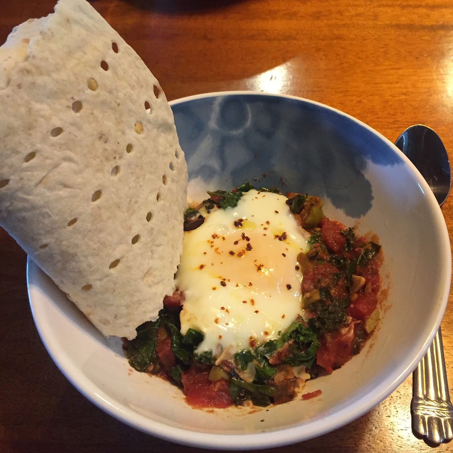 A white and blue bowl with the puttanesca and a poached egg dusted with chili flakes. At the edge of the bowl is a large slice of flatbread.