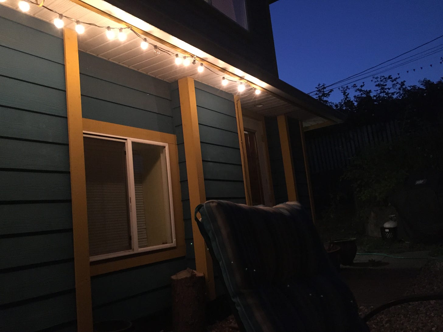 in a dark yard, patio lights create a warm glow from the eaves of a blue and yellow house. in the foreground, a dimly lit chair is visible. in the background, a patch of deep blue evening sky.