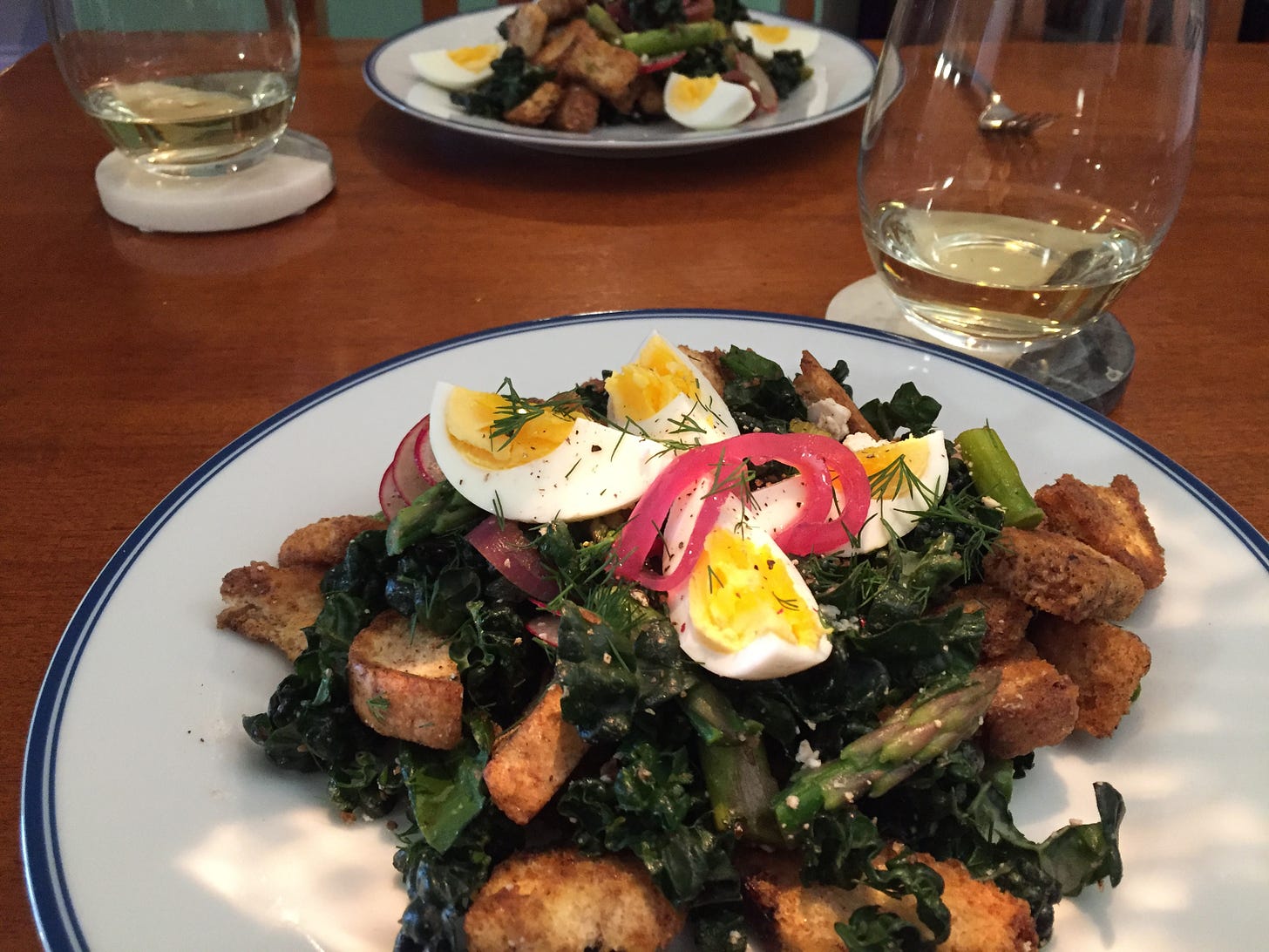A pair of plates sit on a wooden table, each with large kale salad with croutons and asparagus, flecked with dill and bright pink pickled onions, and a boiled egg cut into quarters rests on top. Two stemless glasses of white wine sit on coasters next to the plates.