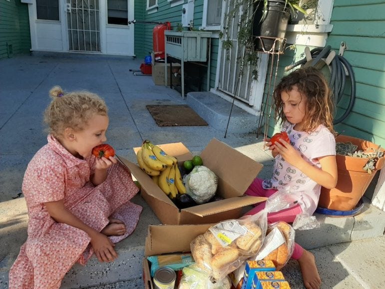 Two young girls sit around a box of food and bite into red tomatoes