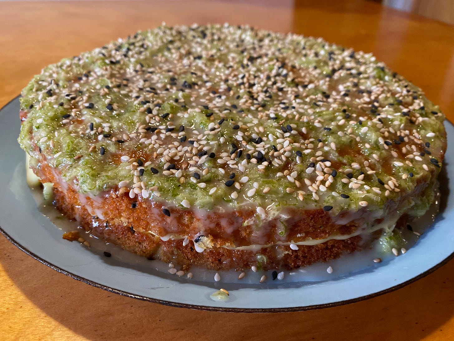 Closeup of a round sandwich cake on a blue plate. The top is covered thin glaze, bright green lime zest, and lots of black and white sesame seeds.