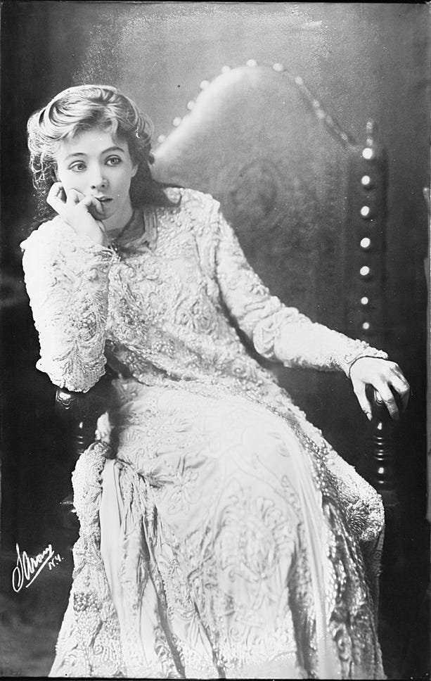a young white woman with loosely curled hair, wearing a white lace gown, sits in an ornate wooden chair. she leans her cheek in one hand and stares at a point beyond the frame with an intense, thoughtful expression. the photograph is from the 19th century and is in black and white.