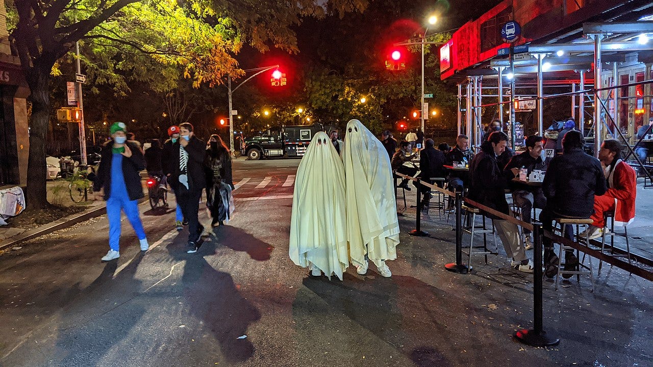 People in costumes celebrating Halloween on a New York street. Two are wearing sheets, with eye holes cut in them, and are costumed as ghosts.