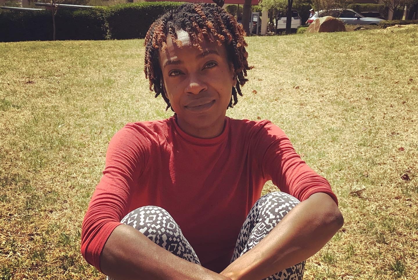 Akilah with two-strand twists, a red shirt, and black and white tights. Seated in grass.