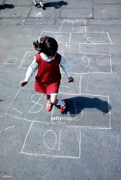 This contains an image of: BRITAIN, A young girl plays hop-scotch, with numbers marked out on...