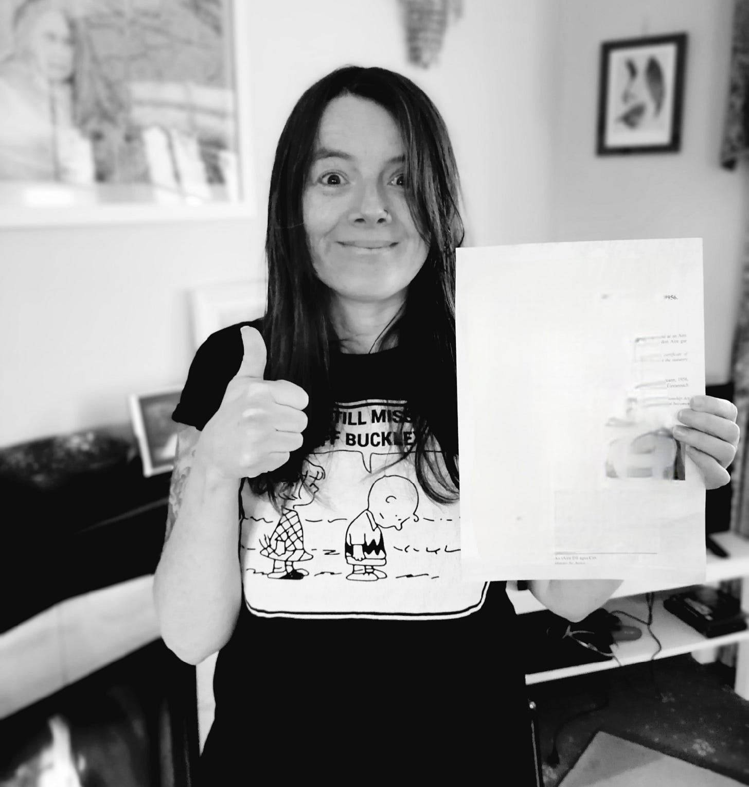 Black and white photo of a smiling woman, holding up a piece of paper in one hand and giving a thumbs up sign with the other. She is in a living room in a house with a large picture on the wall behind her and a lit fire in the fireplace. Black and white photo of a smiling woman, holding up a piece of paper in one hand and giving a thumbs up sign with the other. She is in a living room in a house with a large picture on the wall behind her and a lit fire in the fireplace.