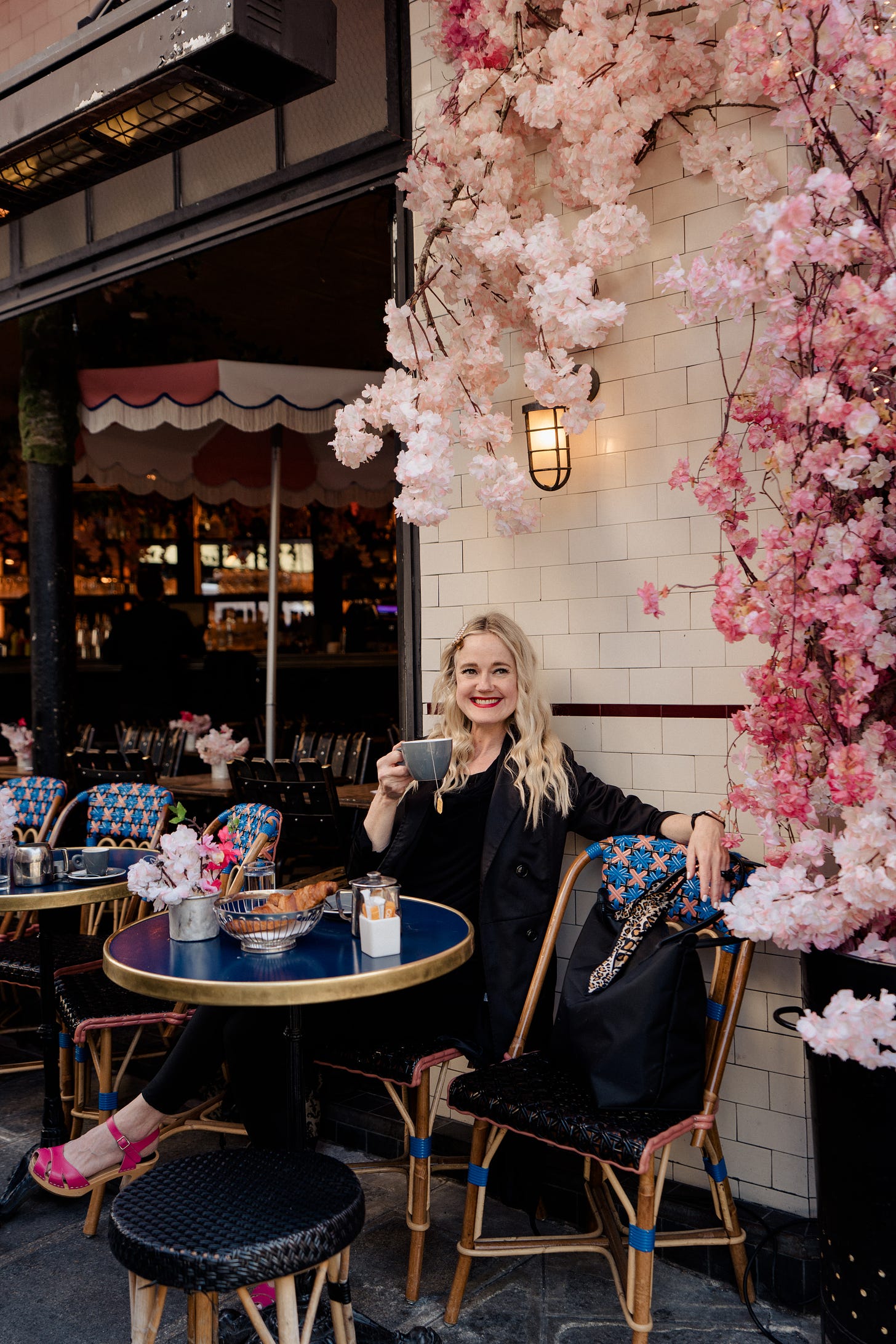 Kimberly, a white woman with long, blonde, wavy hair, red lipstick, black clothes and pink shoes, is sitting at an outdoor cafe table in Paris, holding a coffee and smiling into the camera. Behind her you can see the cafe, along with a tumble of pink blossom. 