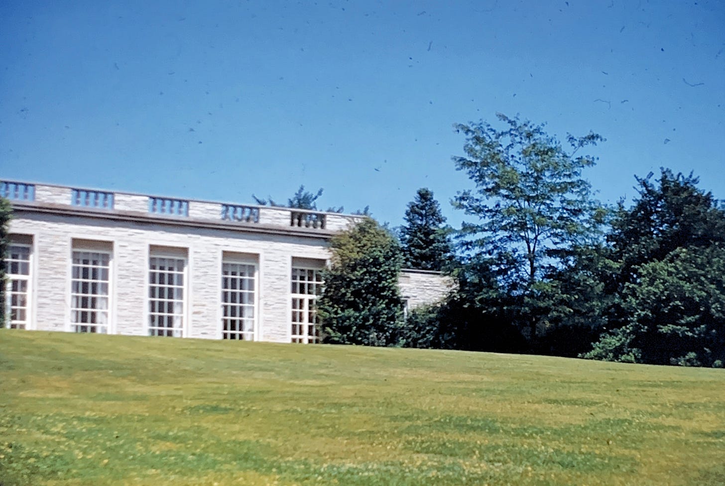 A white stone building with large floor to ceiling windows with trees to the right side and a large green lawn in front.