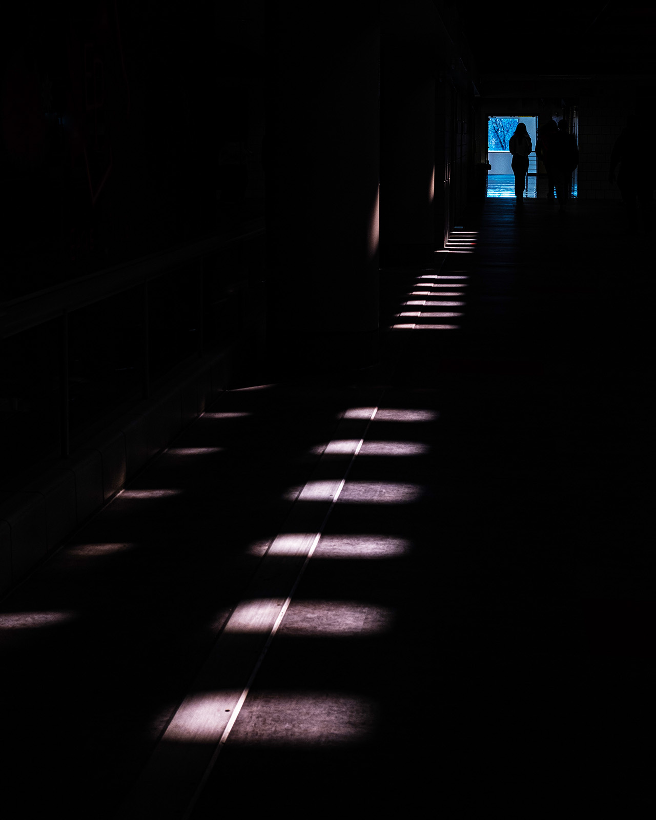 A line of lights illuminates a path on the floor a dark space leading to a person silhouetted in the opening an illuminated hallway.