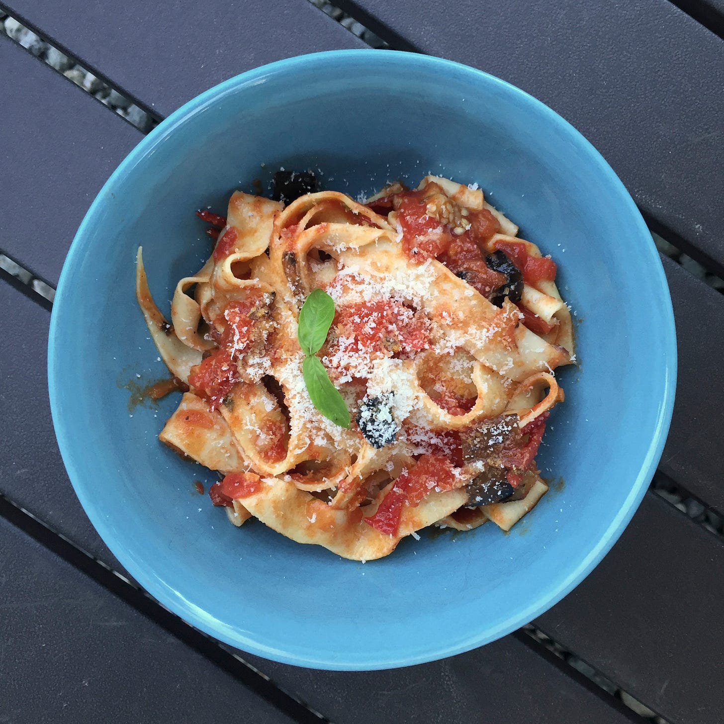 A blue bowl of hand-cut pappardelle in pomodoro sauce, with pieces of grilled eggplant throughout. On top is a dusting of parmesan and two small basil leaves.