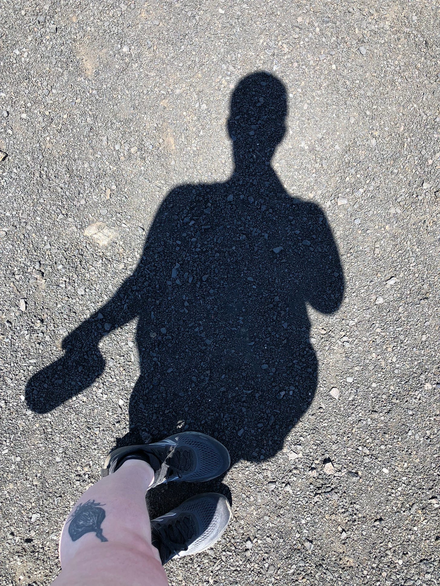 the shadow of a person cast on asphalt with one visible tattooed leg