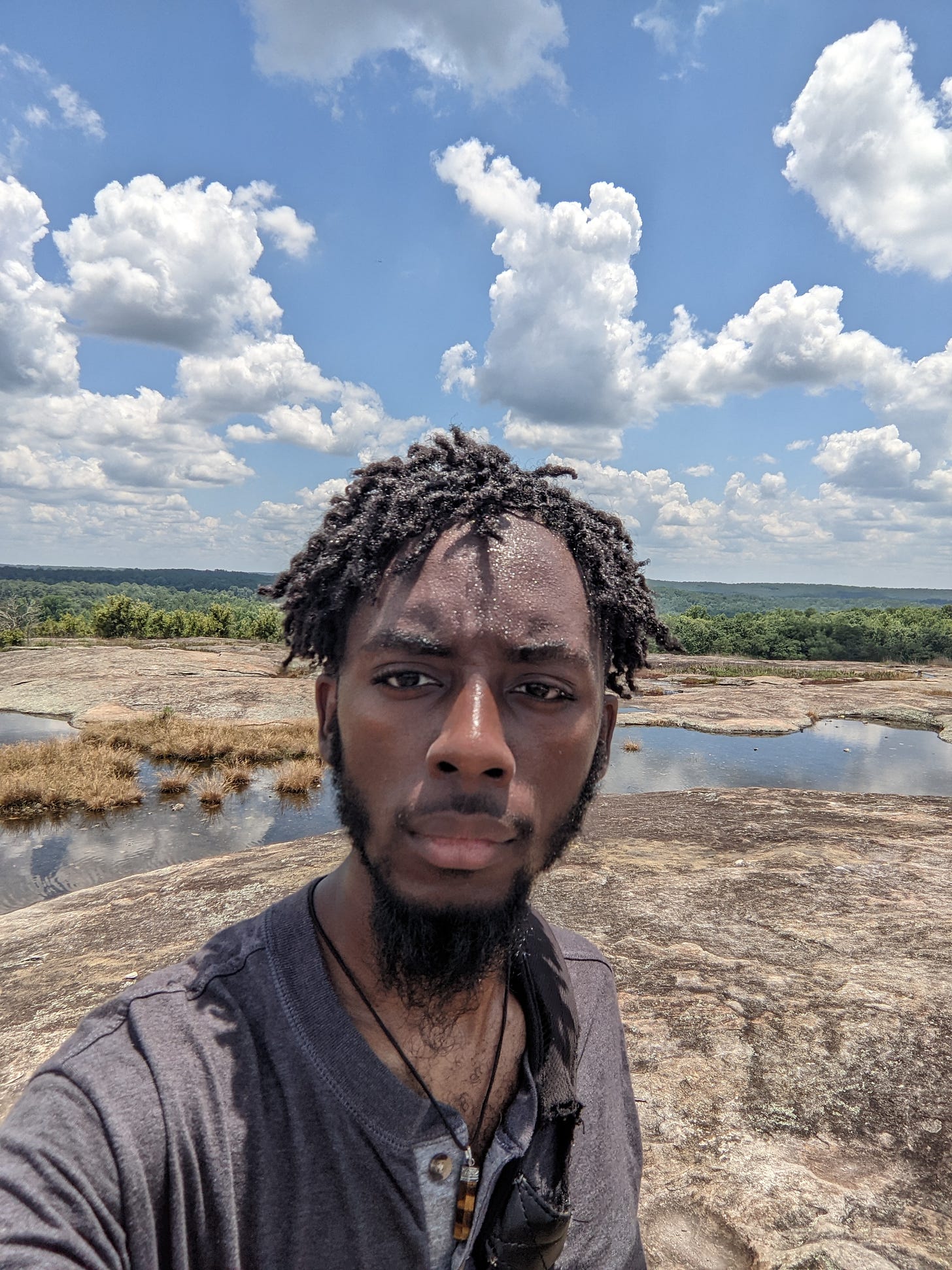 My brown face, sprinkled with sweat, crinkled baby locs atop my head. I'm standing on a so-called mountain with clouds and trees in the distance. There are shallow pools of water behind me on the mountain.