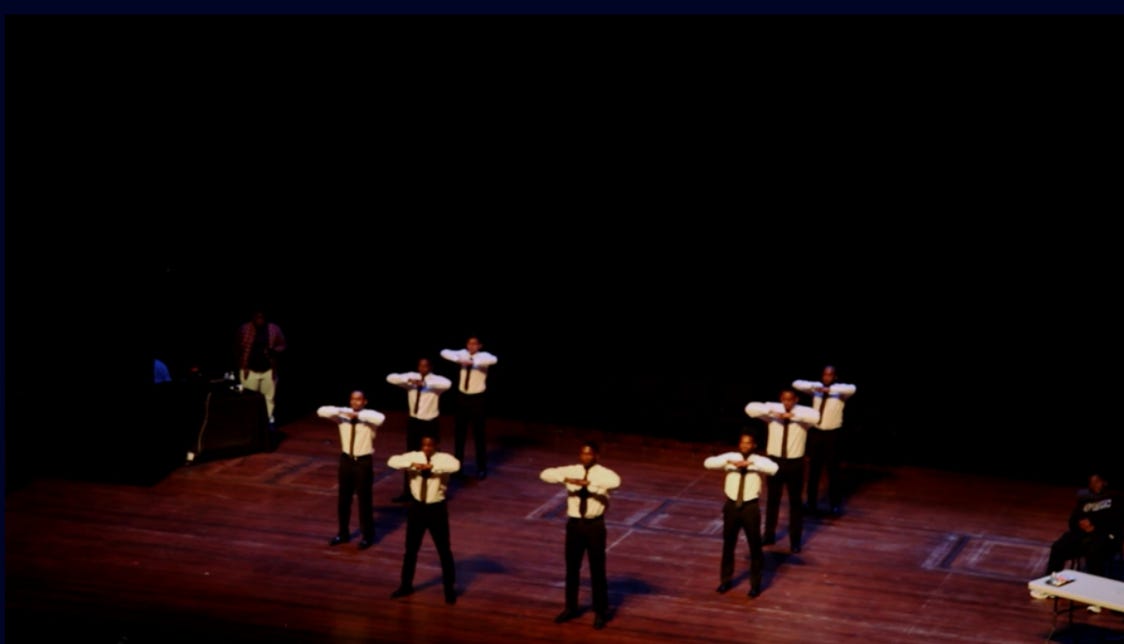 Screenshot of 8 Black men on a stage widely spaced in step formation, all wearing Black pants and ties, with white shirts. 
