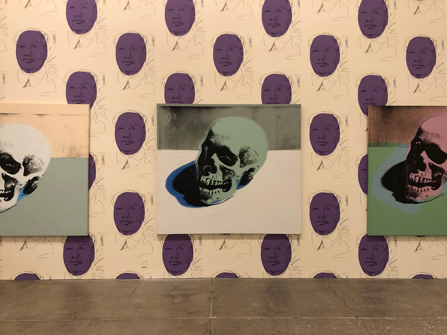 Three large paintings of skulls (white, sea green, and pale pink) are hung on a wall full of faces of Chairman Mao highlighted in purple. The works are by Andy Warhol.
