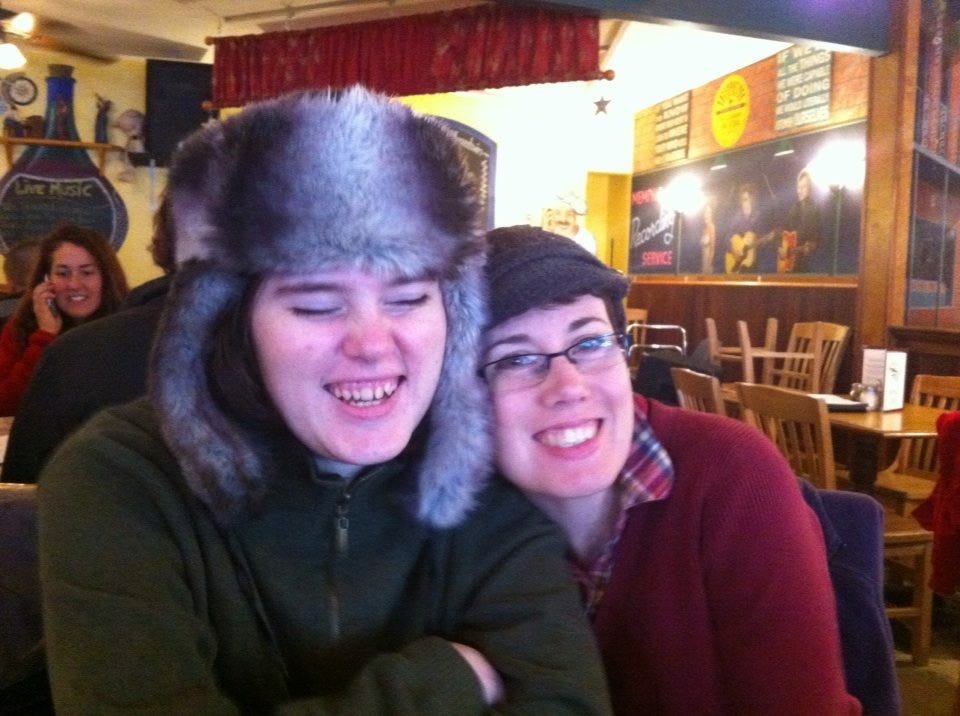 A young white woman is seated on the left, wearing a faux fur earflap hat and green zip-up jacket. Her mother is seated to the right, wearing a burgundy sweater and flannel shirt. Both are smiling at the camera.