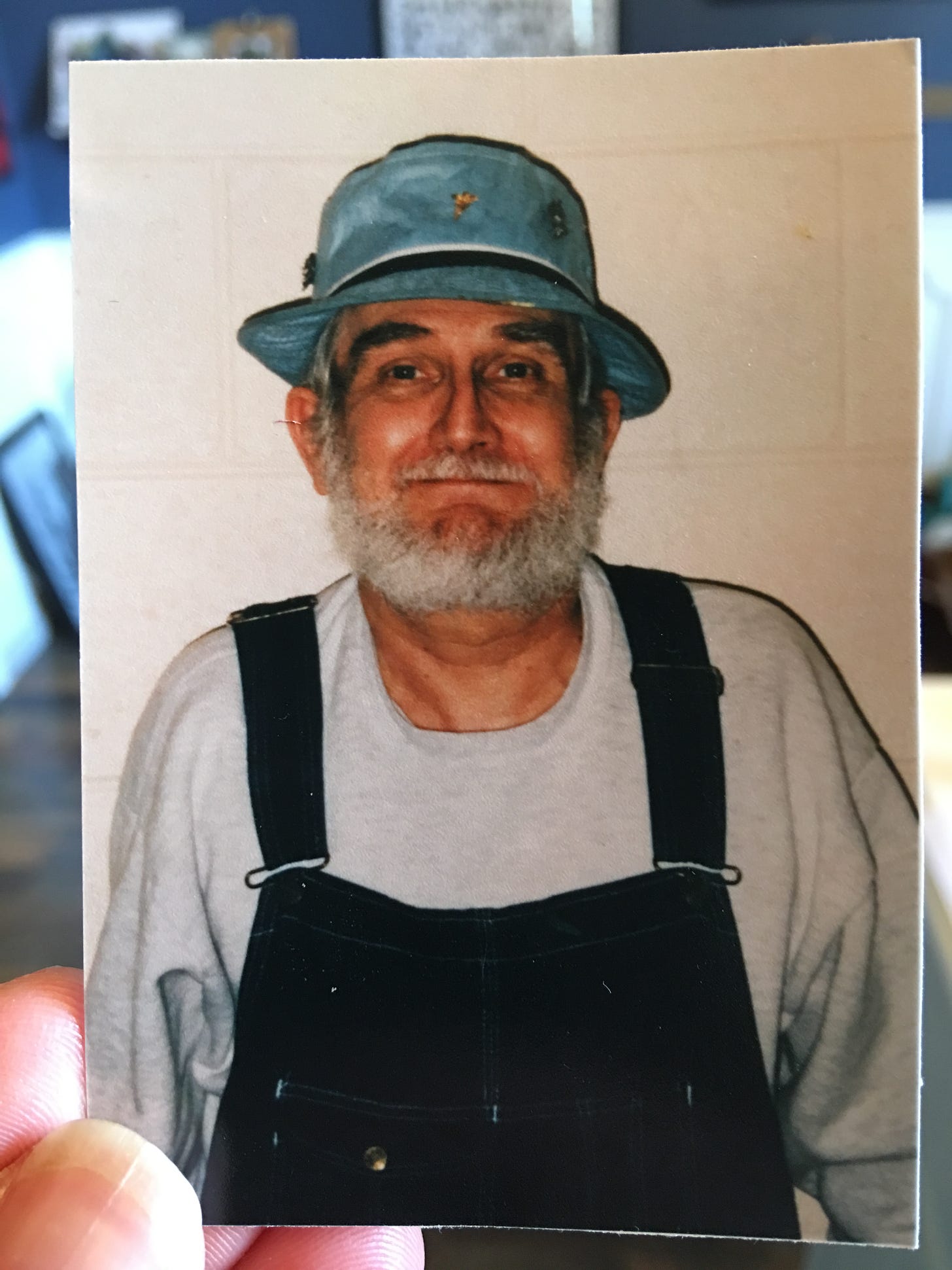 JR stands against a light tan wall, wearing black overalls and a gray sweatshirt. He has a turquoise bucket hat and his smiling lips are closed. He has dark black bushy eyebrows, and interestingly opposite white beard and hair.