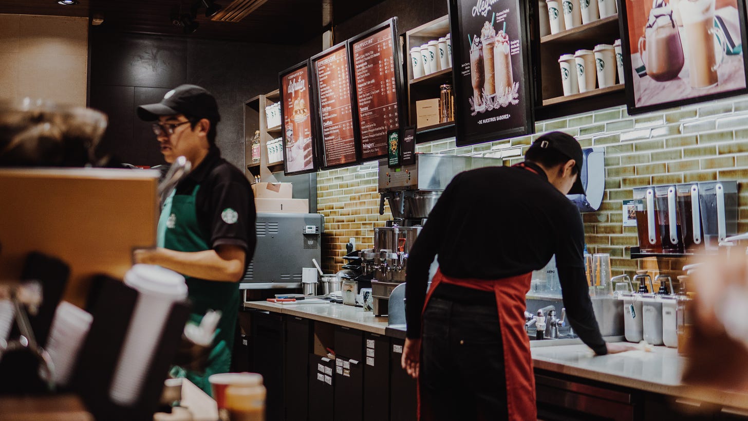 two starbucks baristas in green and red aprons stand behind the espresso bar, one wiping down a counter