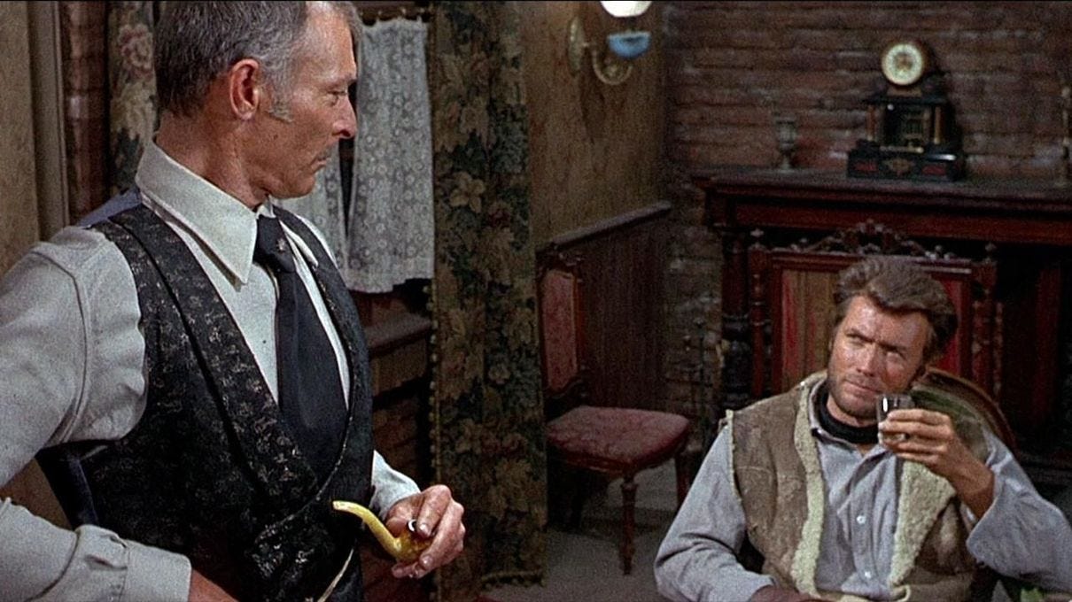 Mortimer stands, frame left, holding his pipe and looking down at a seated Manco, frame right