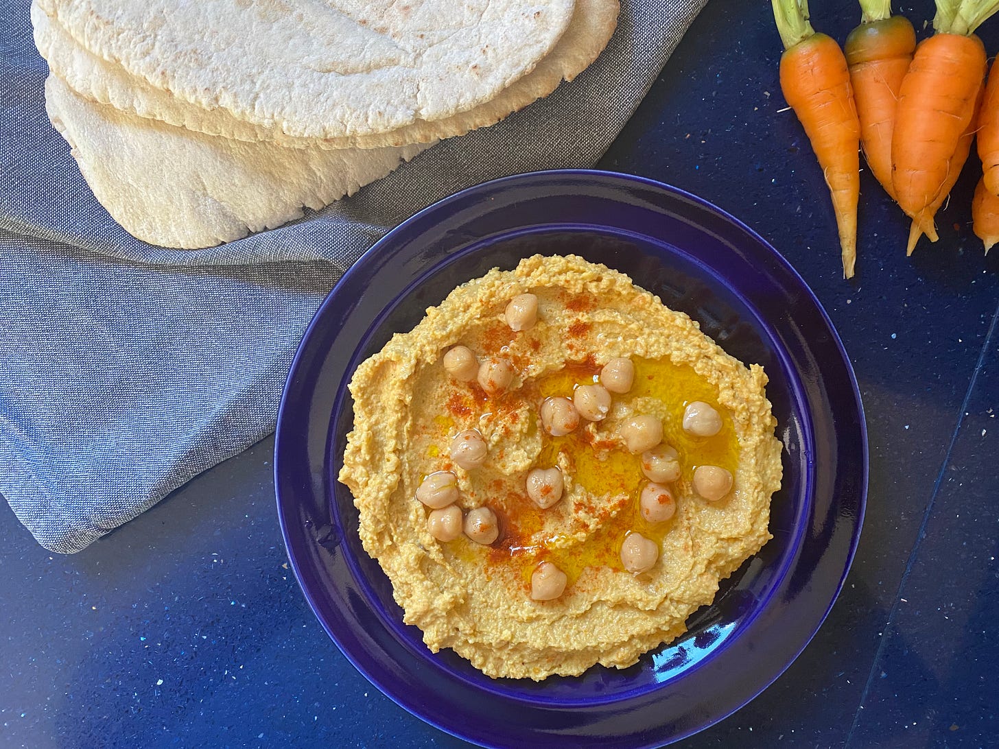 Blue plate with a serving of houmous, scattered with chickpease and paprika. Some pitta breads and carrots are to the side of the plate.