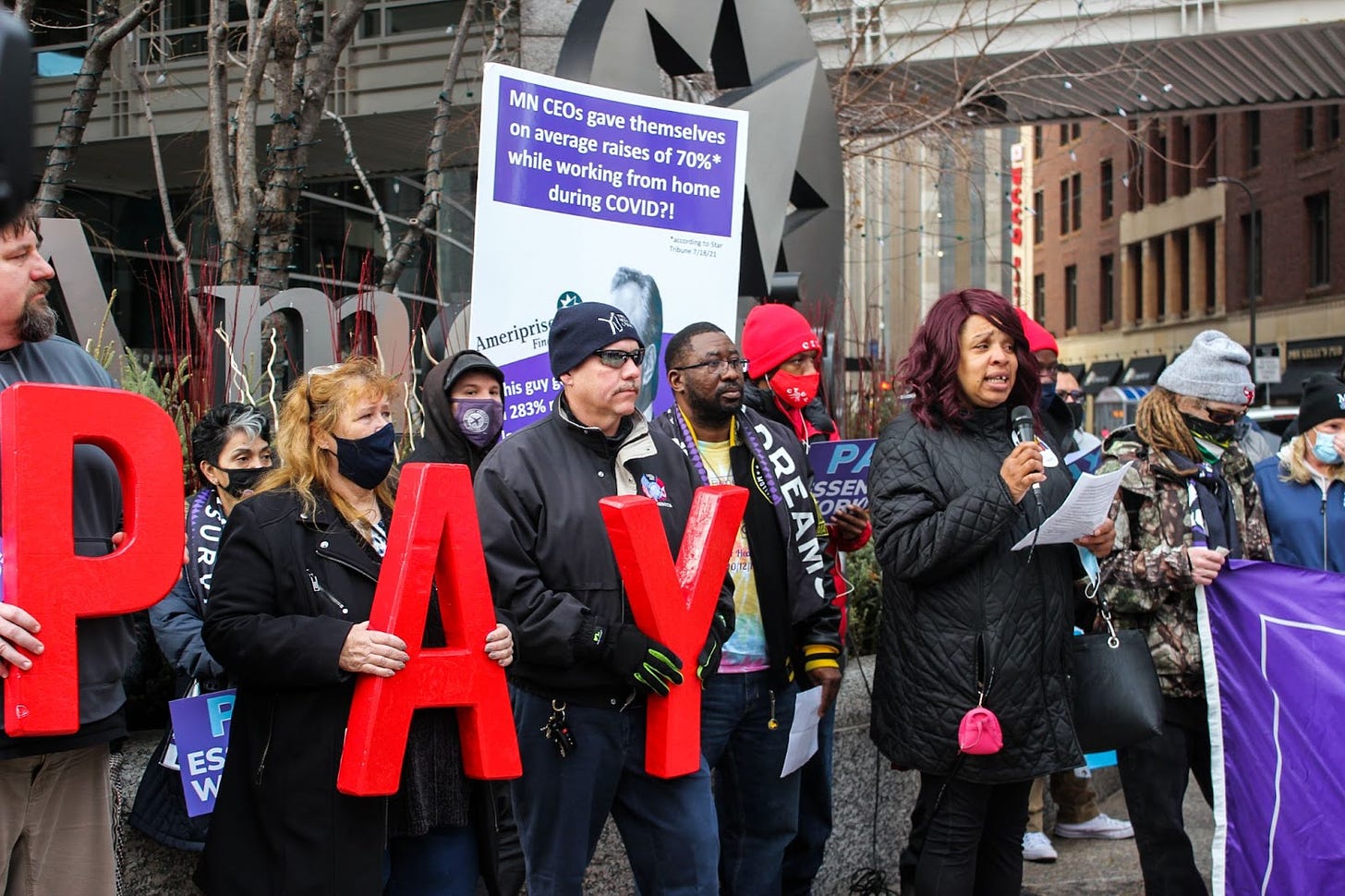members of a crowd hold big red letters spelling "PAY" while a Black woman with red hair stands reading into a microphone