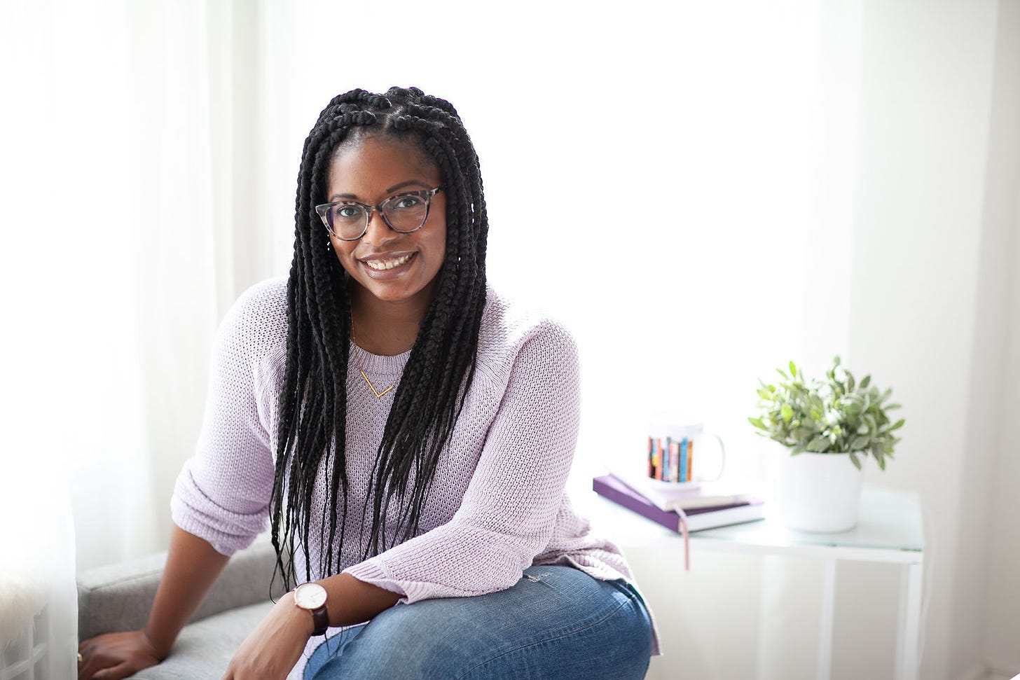 Black woman with box braids, glasses, and a purple sweater sitting facing the camera with a mug and books in the background