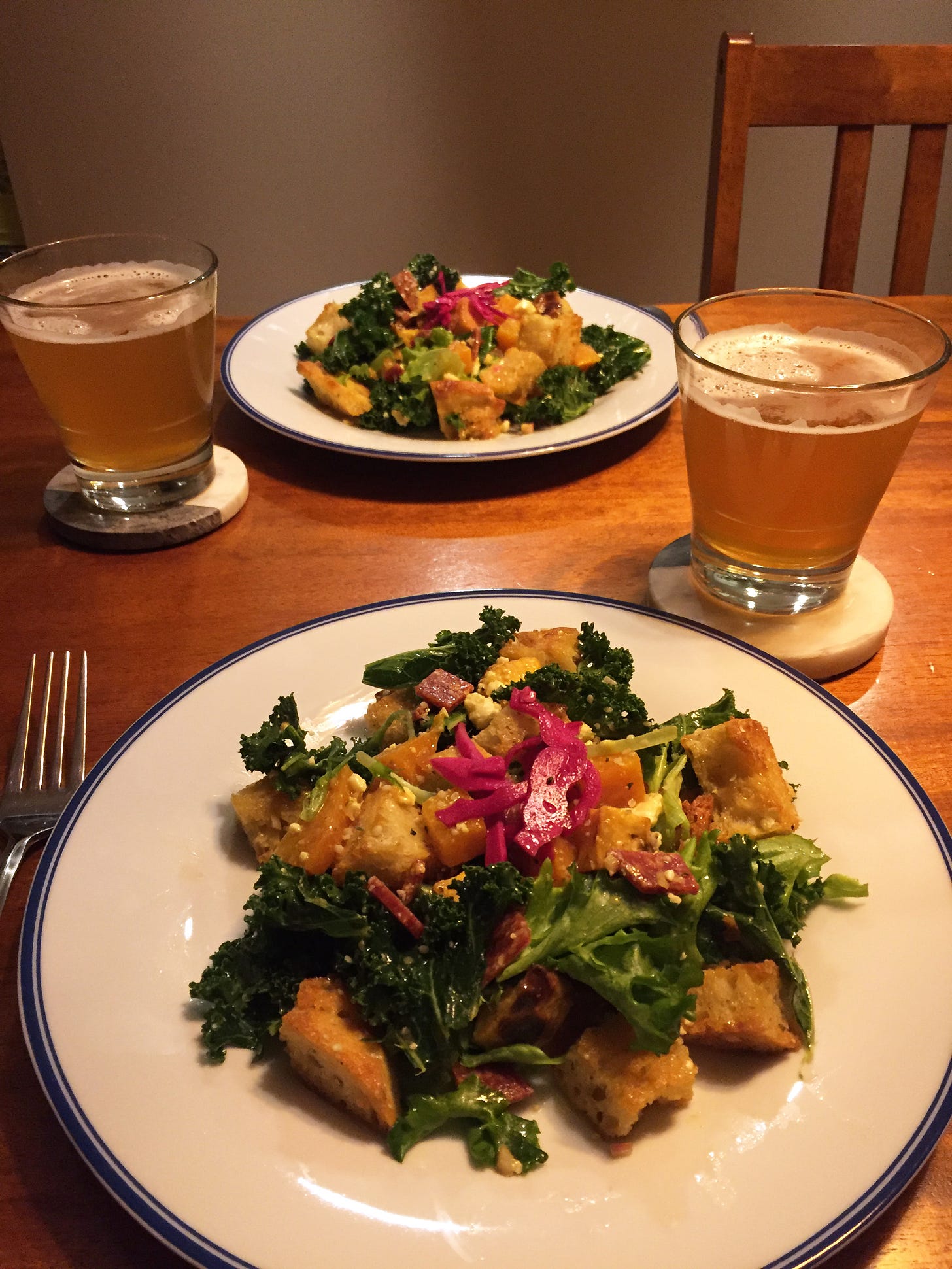 Two plates of panzanella sit across from each other with glasses of beer on coasters next to them. The panzanella is mostly kale and croutons with pieces of golden beet, salami, rutabaga, and pickled cabbage visible throughout.