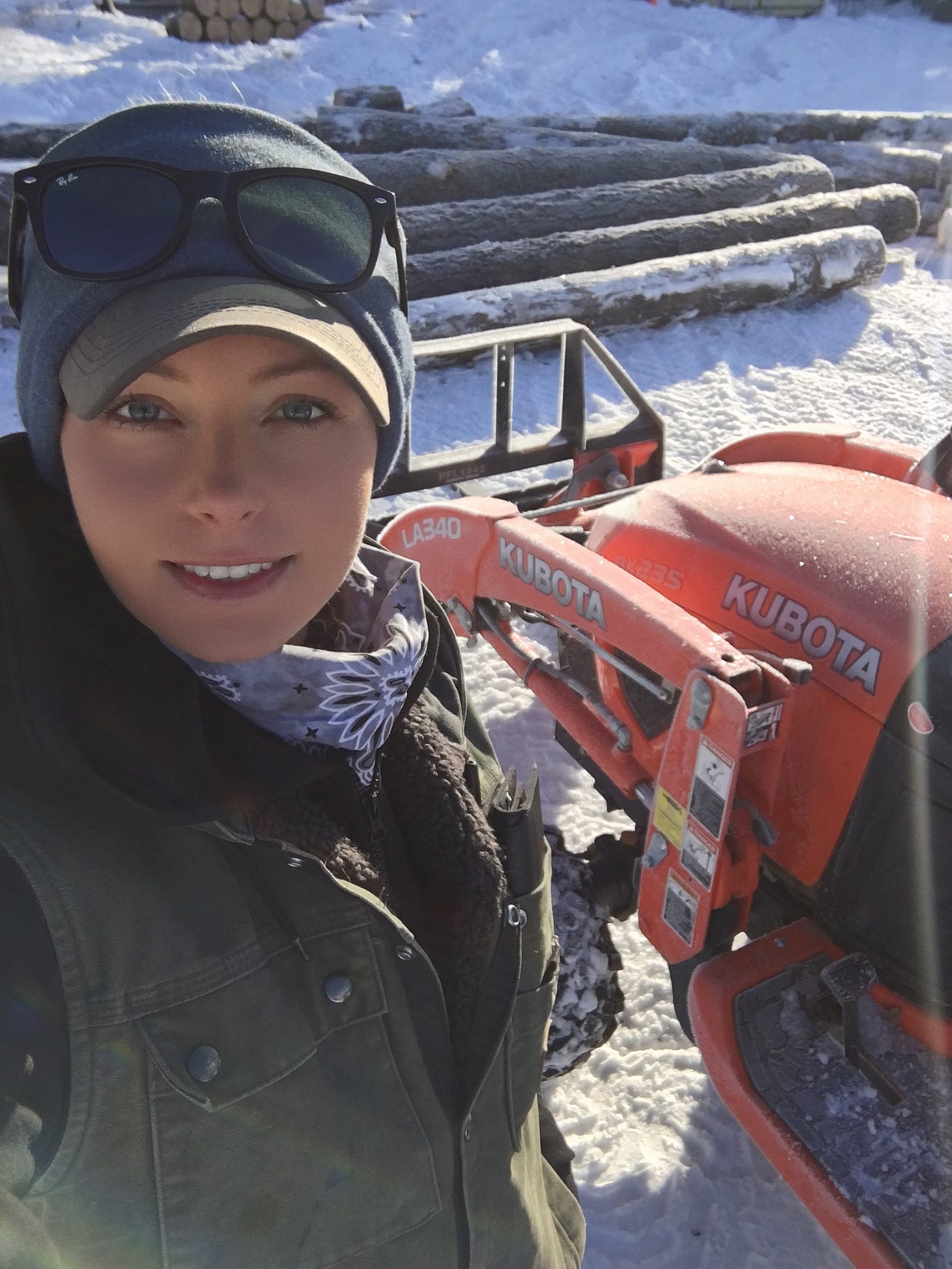 Amy Jay standing in front of a kubota tractor. A pile of logs in the background. 