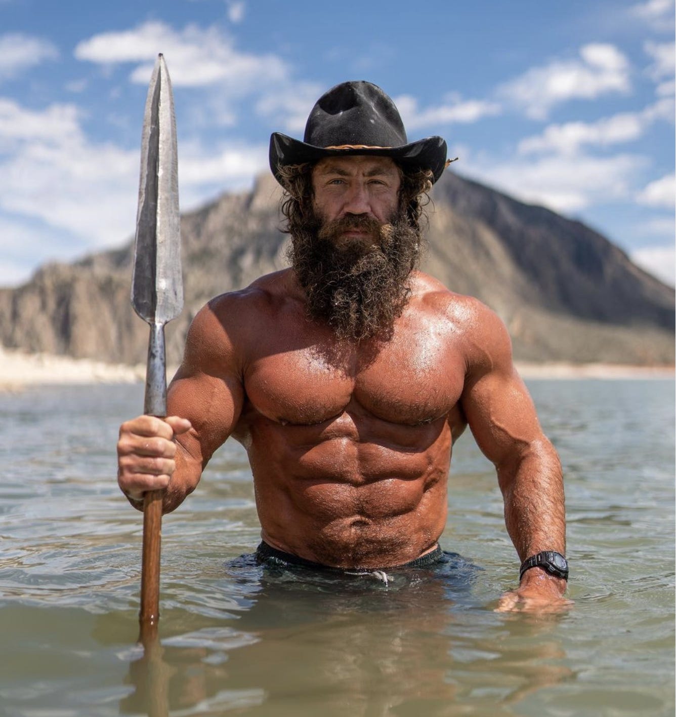 Unnaturally fit man standing in water with a hat holding a spear.