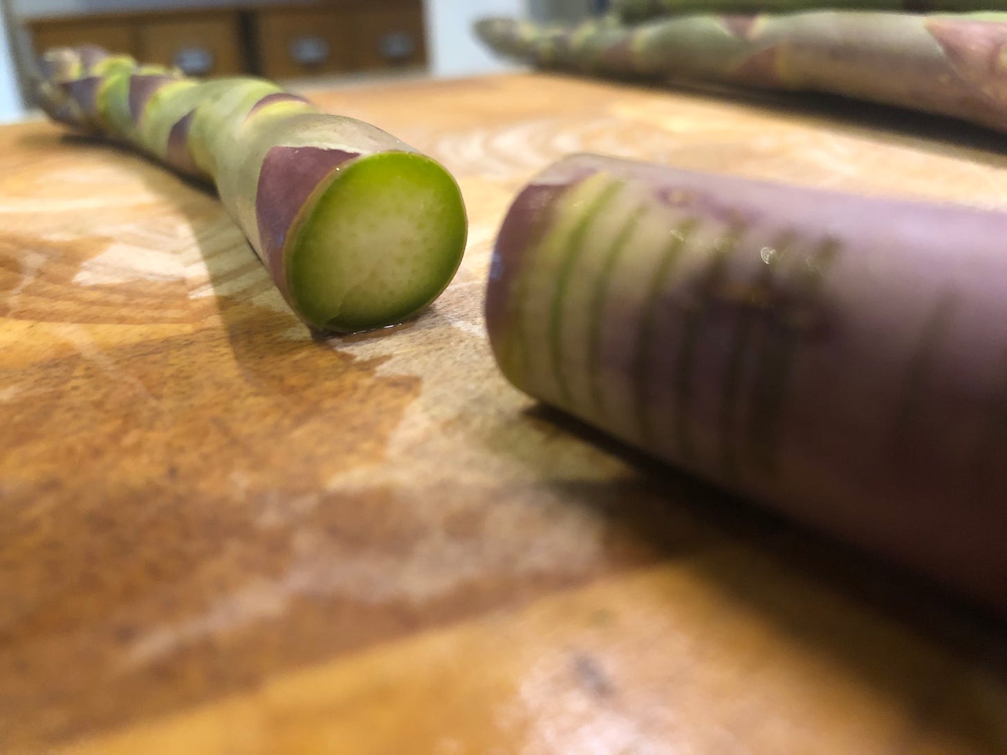 A spear of asparagus on a cutting board. It has been trimmed of its woody end. The woody end has several shallow cuts across it.
