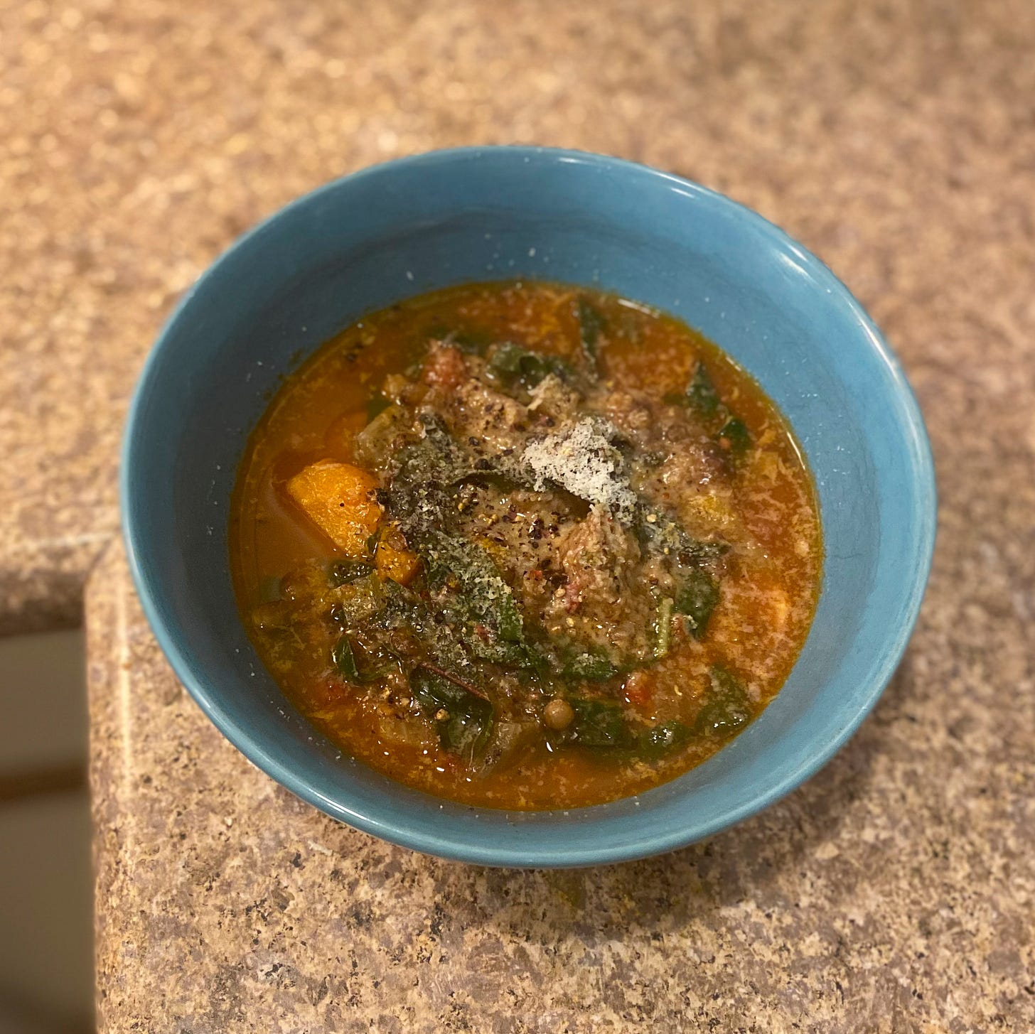 A blue bowl of lentil soup with ribbons of swiss chard, pieces of carrot, and sausage visible. On top is a dusting of grated pecorino.