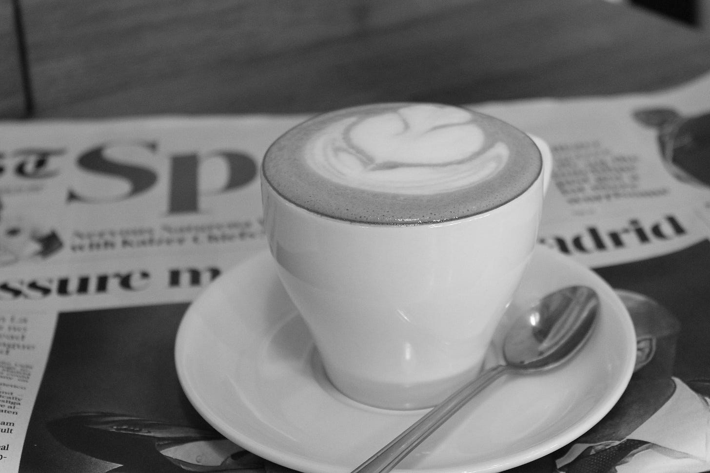 A white coffee cup with latte art and spoon sits atop a newspaper. Via Pixabay.