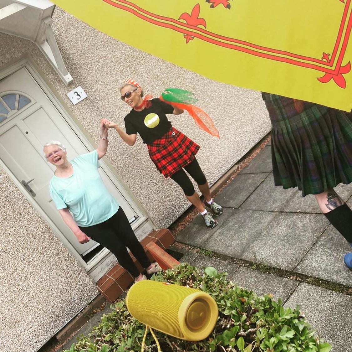 An image of Laura, a middle-aged white woman with short blonde hair, holding hands with an elderly white lady with short white hair. They are standing outside a house with a white door. Laura is wearing a tartan skirt. There is a glimpse of a man wearing a tartan kilt in front but he is wrapped in a yellow Scottish flag with a red border and insignia of lions.