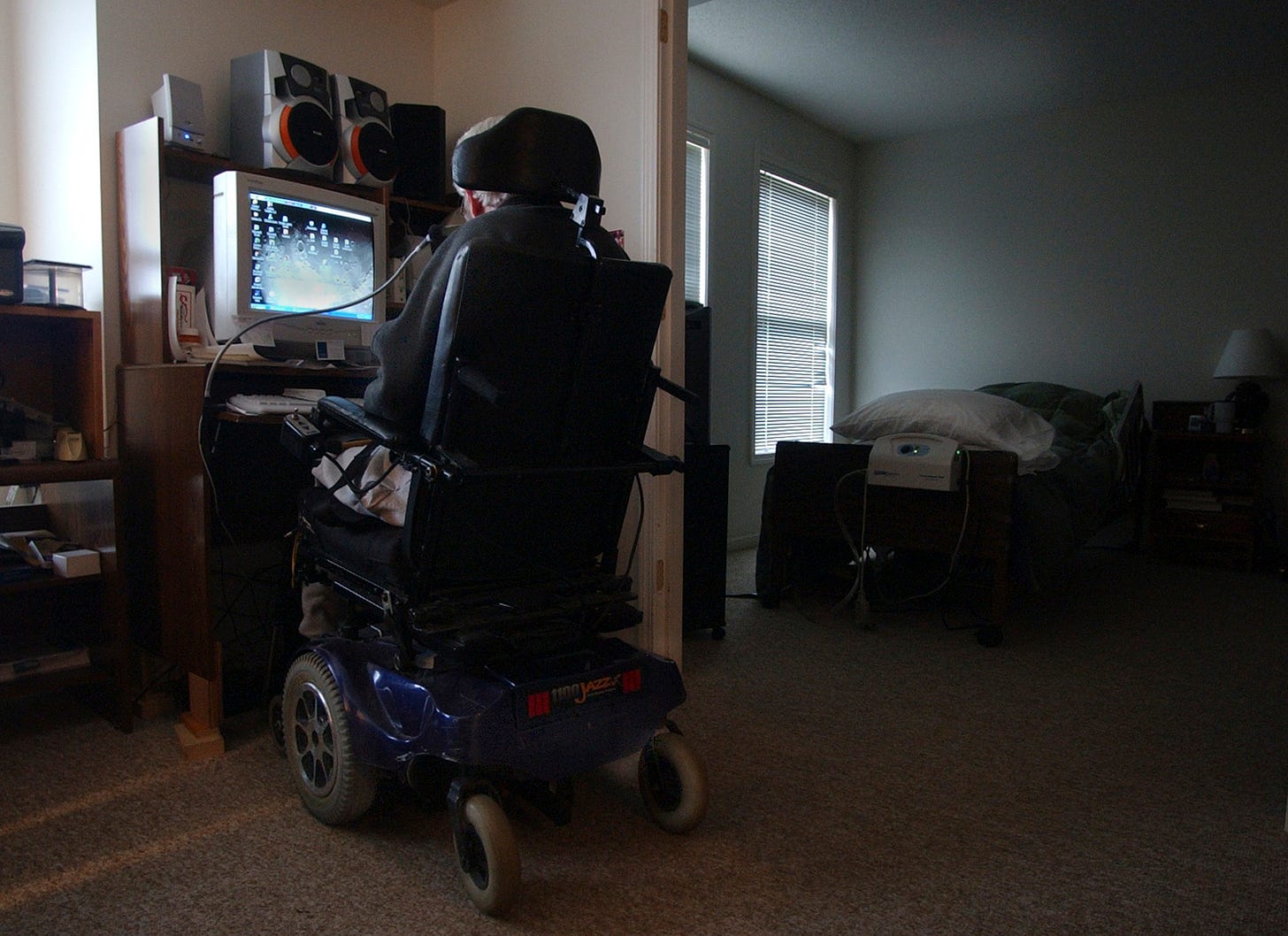 In a dark apartment, we see a motorized wheelchair, the man in it just visible, sitting at a computer.