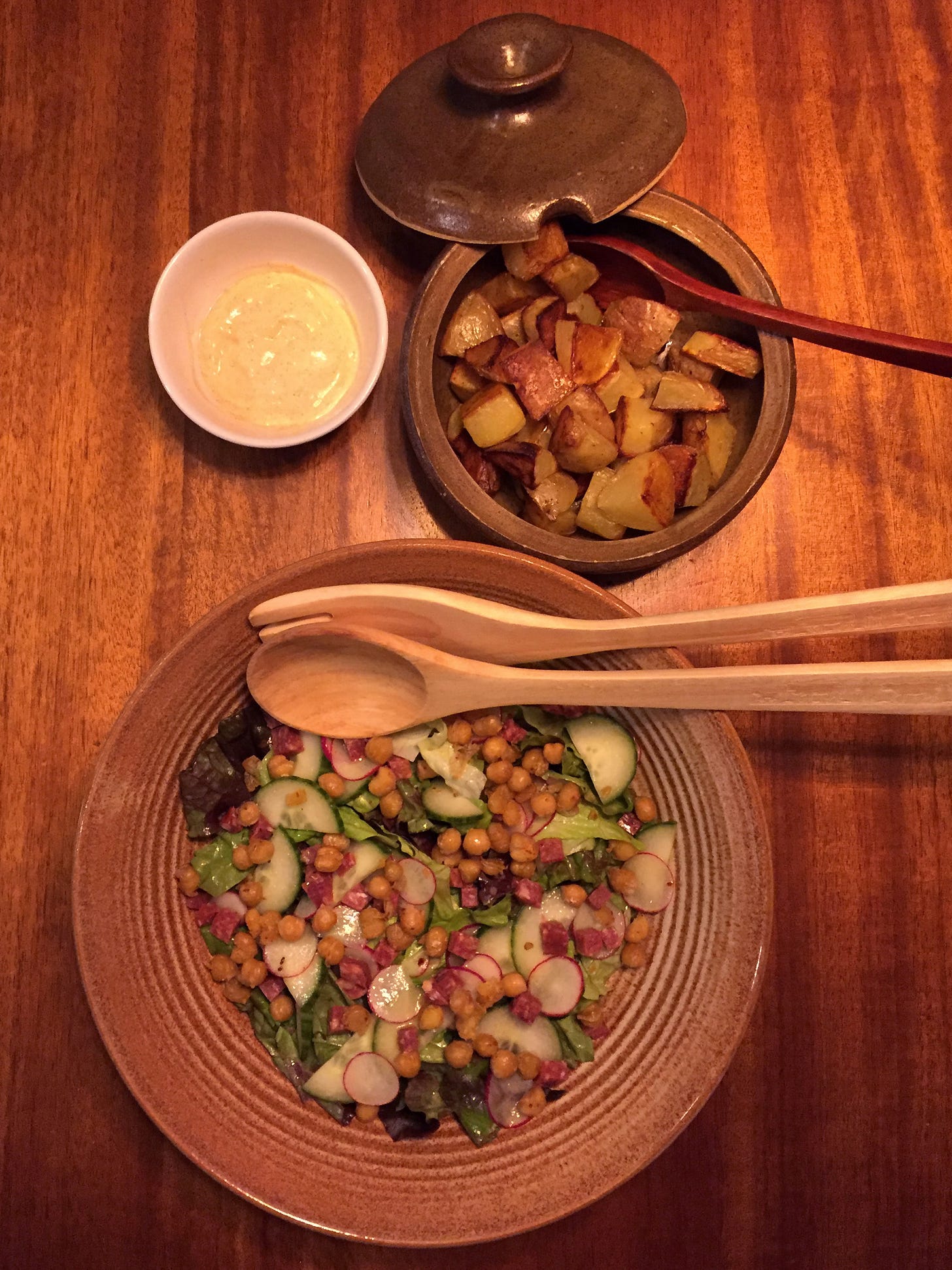 From above, a large ceramic bowl full of green salad with chickpeas and small cubes of salami, with slices of radish and cucumber. Above it is a small tureen filled with browned potatoe pieces, with the lid and a spoon resting at the edge of the dish. Beside this is a small white bowl of the paprika-garlic mayo.