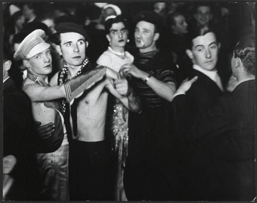 black and white photo of men, some in drag, some shirtless, dancing together